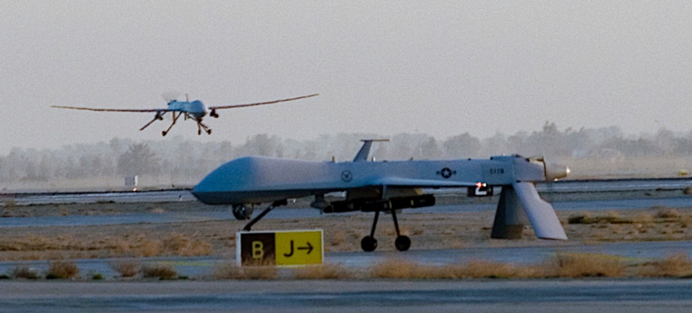 The image shows two unmanned aerial vehicles (UAVs) at what appears to be a military airfield in an arid environment. One UAV is on the ground on a taxiway near a yellow taxiway sign marked 'B' and 'J→', while a second UAV is visible in the background in flight or on approach. The setting appears to be a desert region based on the terrain, sparse vegetation, and hazy atmospheric conditions.