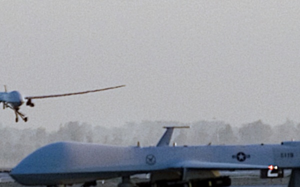 The image shows two unmanned aerial vehicles (UAVs) at what appears to be a military airfield in an arid environment. One UAV is on the ground on a taxiway near a yellow taxiway sign marked 'B' and 'J→', while a second UAV is visible in the background in flight or on approach. The setting appears to be a desert region based on the terrain, sparse vegetation, and hazy atmospheric conditions.