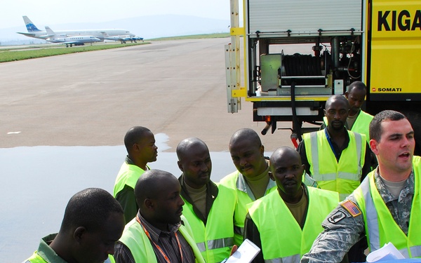 A US Army soldier in military uniform and high-visibility vest is conducting a training or briefing session for airport ground crew workers at Kigali International Airport in Rwanda. The group of approximately 10 workers wearing yellow high-visibility vests are gathered around what appears to be airport ground support equipment, with some taking notes. A commercial aircraft and airport tarmac are visible in the background.