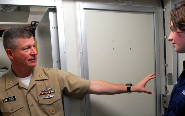 A U.S. Navy officer in khaki service uniform is speaking with a younger person wearing a dark blue U.S. Coast Guard uniform. The Navy officer is gesturing with an outstretched arm toward the Coast Guard member, appearing to explain or direct something. The setting appears to be the interior of a naval vessel or military facility.