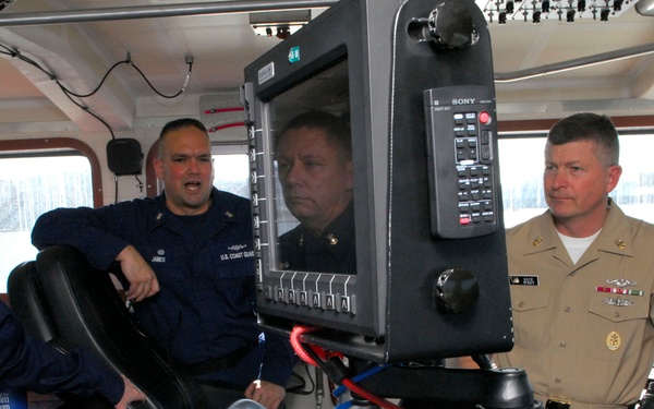 Four military personnel are gathered on the bridge/wheelhouse of a vessel, appearing to discuss or review navigation equipment. Two individuals are wearing dark blue U.S. Coast Guard uniforms, one is in tan/khaki Navy uniform with ribbons and insignia, and one appears to be in a darker service uniform. They are gathered around a Sony navigation/radar display monitor mounted on the vessel's helm console.