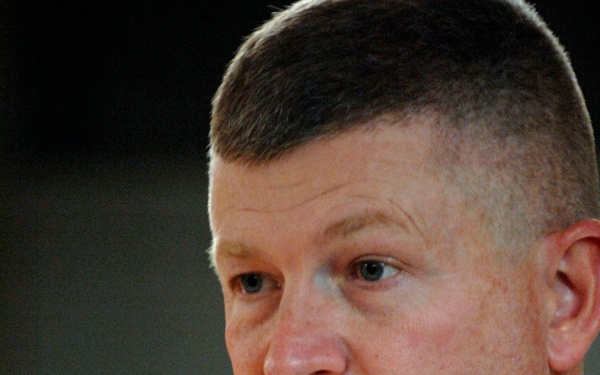 A middle-aged male in a tan/khaki military uniform is speaking, with his mouth open mid-sentence. A gold insignia pin is visible on his collar. In the foreground, partially out of focus, are hands belonging to another individual wearing a dark wristwatch.