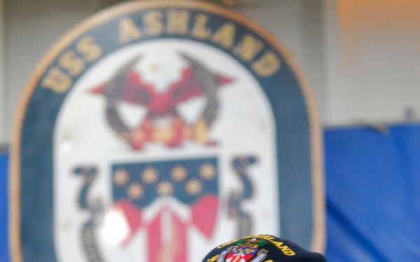 A military officer wearing a dark uniform and a USS Ashland cap is photographed aboard the USS Ashland (LSD-48), a U.S. Navy dock landing ship. The large official ship's crest/seal is prominently displayed in the background. The officer appears to be speaking or giving remarks in what looks like a hangar or interior deck space.
