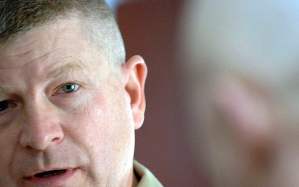 A middle-aged male military officer in a tan/khaki uniform is engaged in conversation with another person whose back is partially visible in the foreground. The officer appears to be speaking, with his mouth slightly open. The image is a close-up portrait-style photograph taken in an indoor setting.