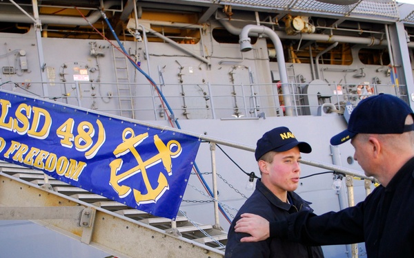 Three U.S. Navy personnel are standing on a pier next to a naval vessel identified as USS Ashland (LSD 48). A banner on the gangway reads 'ASHLAND (LSD 48) - LIBERTY DEFEND FREEDOM' with an anchor emblem in gold on a blue background. One sailor appears to be receiving guidance or instruction from a senior officer who has his hand on the sailor's shoulder.