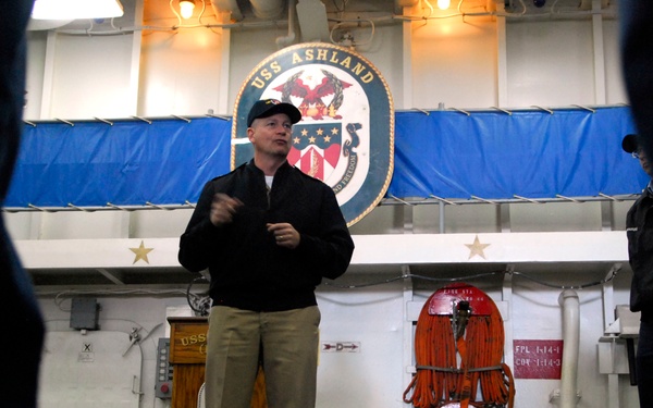 A man in a black jacket and khaki pants stands and speaks aboard the USS Ashland, a U.S. Navy vessel. He appears to be addressing an audience, with listeners visible in the foreground as silhouettes. The ship's official seal/crest is prominently displayed on the wall behind him, along with blue banners and orange fire hoses.