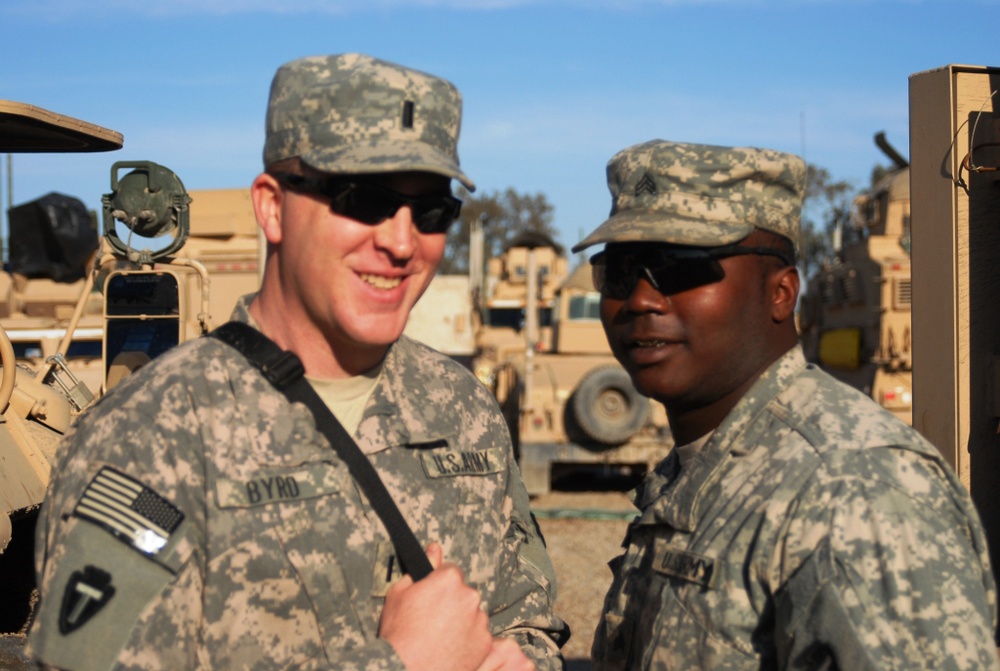 Two U.S. Army soldiers in ACU (Army Combat Uniform) digital camouflage are posing together outdoors at what appears to be a forward operating base or motor pool. Both wear patrol caps and sunglasses, with military vehicles visible in the background. The soldier on the left has the name tape 'BYRD' and a U.S. Army tape visible on his uniform.