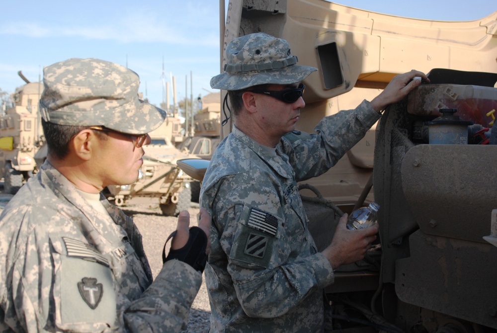 Two U.S. Army soldiers in ACU (Army Combat Uniform) digital camouflage are performing maintenance or inspection on a military vehicle, likely a HMMWV or MRAP, in what appears to be a forward operating base motor pool. Multiple military vehicles are visible in the background, suggesting a staging or maintenance area. The setting and uniforms indicate a deployment environment, likely in the Middle East based on the terrain and vehicle types.