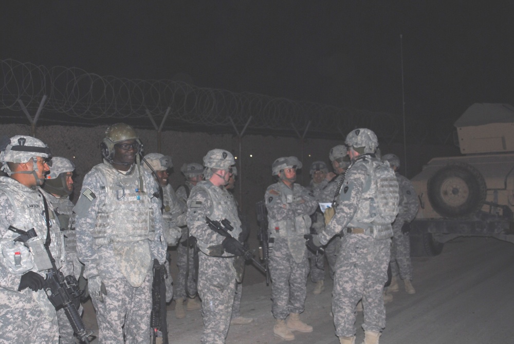 A group of U.S. Army soldiers in full combat gear are assembled outdoors at night, appearing to receive a briefing or conduct a pre-mission formation. They are wearing Army Combat Uniforms (ACU) with helmets, body armor, and carrying weapons. The setting appears to be a forward operating base or secured perimeter, indicated by concertina wire visible in the background.