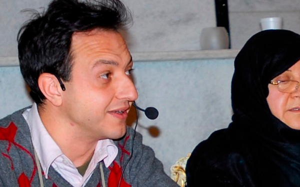 Two individuals are seated at a table with a red tablecloth, appearing to be participants in a panel discussion or conference. The man on the left is wearing a headset microphone and speaking, while the woman on the right, wearing a black hijab and glasses, appears to be listening. Documents, a water bottle, and a foam cup are visible on the table in front of them.