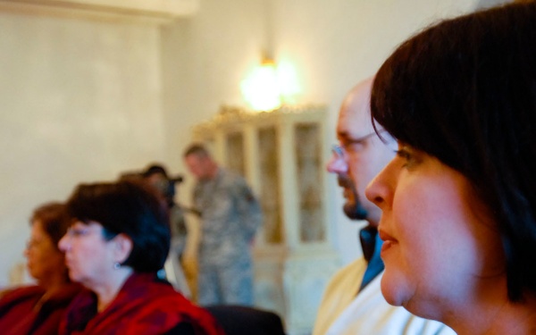 A group of people are seated at tables in what appears to be a meeting or briefing room, attentively facing a presenter or speaker out of frame. The attendees include both military personnel in camouflage uniforms and civilians in everyday clothing. Colored documents and folders are visible on the tables in front of them.