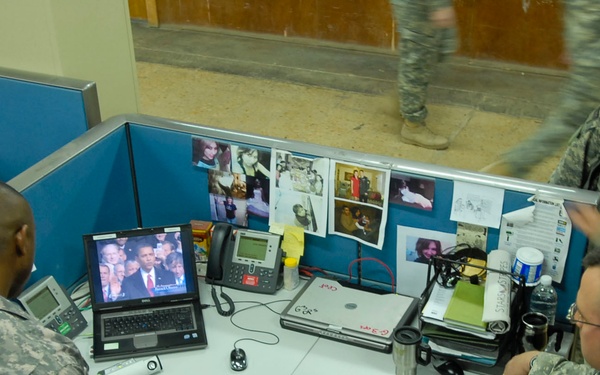 Several U.S. Army soldiers in ACU (Army Combat Uniform) camouflage are gathered around a cubicle workstation watching a broadcast on a Dell laptop computer. The setting appears to be a military office or operations center, with standard office cubicle partitions, phones, and personal photos pinned to the dividers. The soldiers appear to be attentively watching what appears to be a significant televised event.