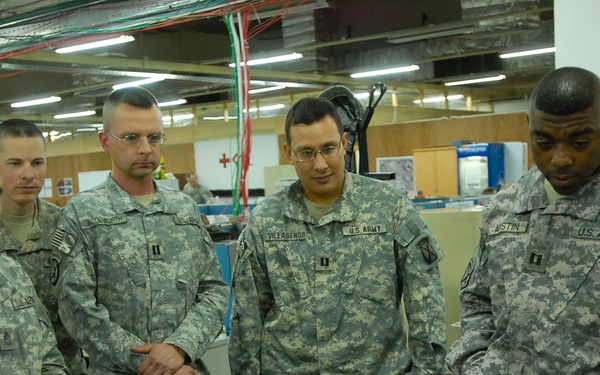 Five U.S. Army soldiers in ACU (Army Combat Uniform) camouflage are gathered around a workstation in what appears to be a military operations center or command post. They appear to be reviewing information on a monitor or display surface. The setting is an indoor facility with overhead fluorescent lighting and visible cable management infrastructure.