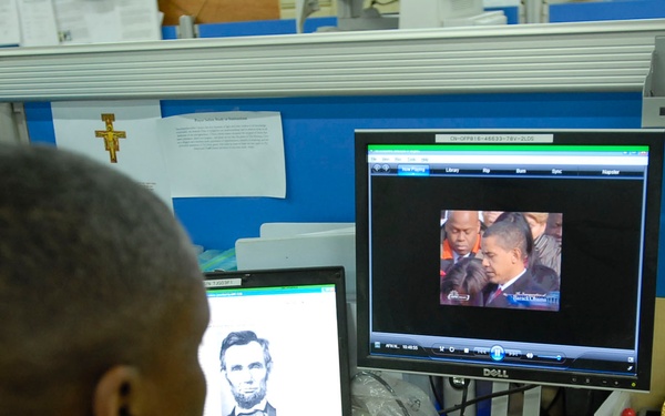 A person in military camouflage uniform sits at a cubicle desk viewing two computer screens. The laptop displays a black-and-white portrait illustration, while the Dell monitor shows a video stream with a caption reading 'Barack H. Obama'. The workspace includes religious items, a CompTIA security certification, and various desk accessories.