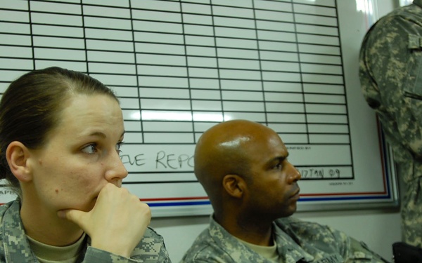 Three U.S. Army soldiers in Army Combat Uniform (ACU) digital camouflage pattern are seated or standing in what appears to be a briefing or administrative room. All soldiers wear the 10th Mountain Division patch ('Mountain' tab with crossed rifles insignia) on their left shoulders, along with reversed U.S. flag patches. They appear to be attending a briefing or waiting in a military administrative setting.