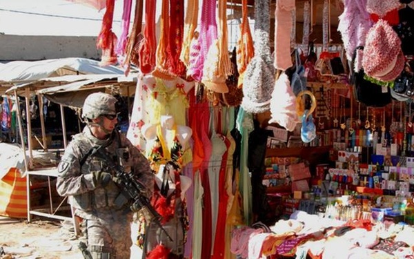Two U.S. military soldiers in combat gear and ACU (Army Combat Uniform) are patrolling through an open-air market bazaar in what appears to be Iraq or Afghanistan. The market stalls display a wide variety of colorful goods including clothing, accessories, and consumer products. A red banner with Arabic script is visible in the background.