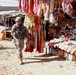 Two U.S. military soldiers in combat gear and ACU (Army Combat Uniform) are patrolling through an open-air market bazaar in what appears to be Iraq or Afghanistan. The market stalls display a wide variety of colorful goods including clothing, accessories, and consumer products. A red banner with Arabic script is visible in the background.