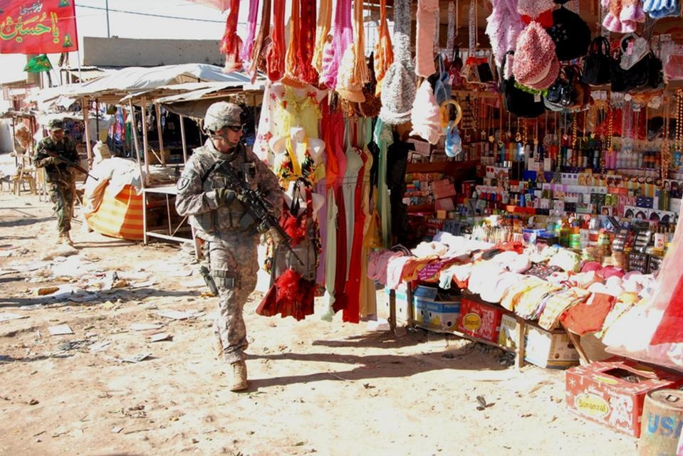Two U.S. military soldiers in combat gear and ACU (Army Combat Uniform) are patrolling through an open-air market bazaar in what appears to be Iraq or Afghanistan. The market stalls display a wide variety of colorful goods including clothing, accessories, and consumer products. A red banner with Arabic script is visible in the background.