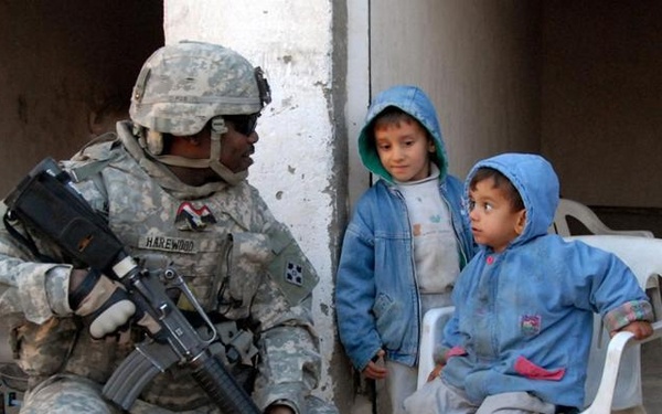 A U.S. Army soldier, identified by name tape as 'HAREWOOD,' crouches down to interact with two young children near a concrete pillar. The soldier is in full combat gear including ACU (Army Combat Uniform) and is armed with an M4 carbine. The children, wearing blue hooded jackets, appear calm — one is seated on a white plastic chair.