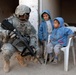 A U.S. Army soldier, identified by name tape as 'HAREWOOD,' crouches down to interact with two young children near a concrete pillar. The soldier is in full combat gear including ACU (Army Combat Uniform) and is armed with an M4 carbine. The children, wearing blue hooded jackets, appear calm — one is seated on a white plastic chair.