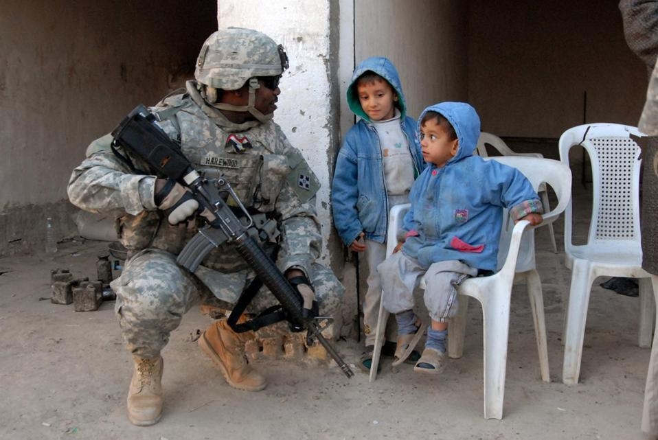 A U.S. Army soldier, identified by name tape as 'HAREWOOD,' crouches down to interact with two young children near a concrete pillar. The soldier is in full combat gear including ACU (Army Combat Uniform) and is armed with an M4 carbine. The children, wearing blue hooded jackets, appear calm — one is seated on a white plastic chair.