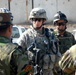 The image shows a group of military personnel in a outdoor setting, appearing to be in a briefing or coordination meeting. A U.S. Army soldier wearing ACU (Army Combat Uniform) with the name tape 'WILLIAMS' is prominently visible in the center, interacting with what appear to be local or allied forces. The setting suggests a forward operating base or checkpoint environment in a Middle Eastern location.