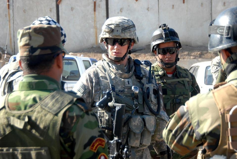 The image shows a group of military personnel in a outdoor setting, appearing to be in a briefing or coordination meeting. A U.S. Army soldier wearing ACU (Army Combat Uniform) with the name tape 'WILLIAMS' is prominently visible in the center, interacting with what appear to be local or allied forces. The setting suggests a forward operating base or checkpoint environment in a Middle Eastern location.