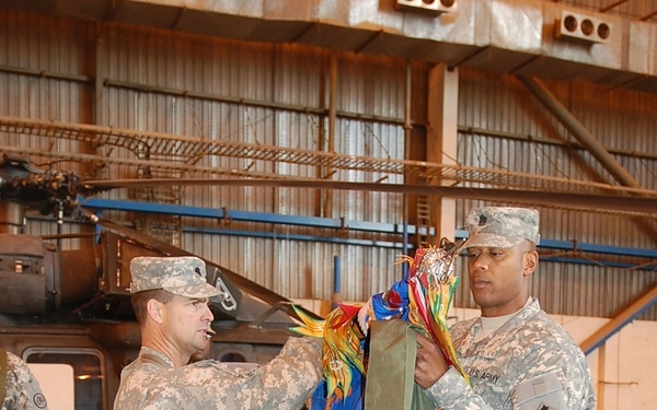 Two U.S. Army soldiers in ACU (Army Combat Uniform) digital camouflage are performing a military colors ceremony inside an aircraft hangar. They are handling what appears to be a unit guidon or colors with multiple streamers, likely during a casing or uncasing ceremony. A military helicopter, consistent with a UH-60 Black Hawk, is visible in the background.