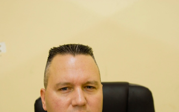 A U.S. Army soldier is seated in a black leather chair against a light-colored wall. The soldier is wearing Army Combat Uniform (ACU) with visible name tape reading 'KASCH' and a 'U.S. AR[MY]' branch tape. A rank insignia consistent with Sergeant First Class or Master Sergeant is visible on the chest pocket.