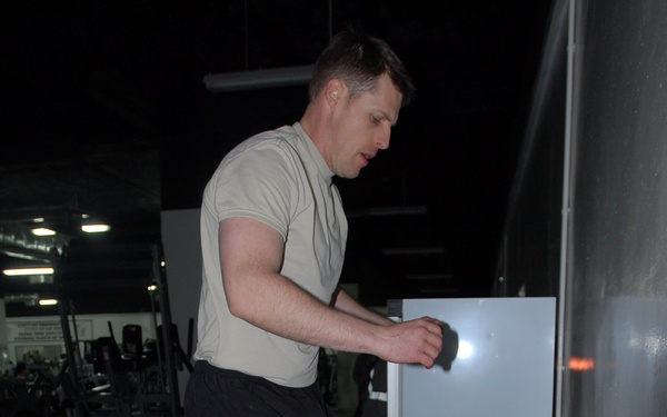 A man is performing a step-up exercise on a black plyometric box in what appears to be a gym or fitness facility. He is wearing a light tan t-shirt, black shorts, and gray athletic shoes, with one knee raised in mid-exercise. The background shows gym equipment and weight racks in a dimly lit space.