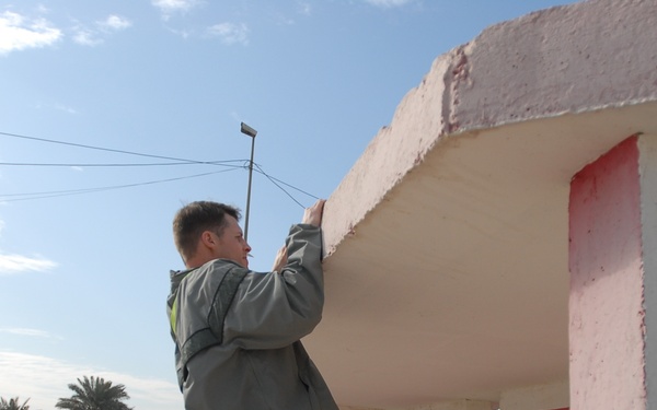 A young man in casual athletic wear (gray hoodie and black shorts) is performing a pull-up or hanging exercise on the ledge of a building with pink and white stucco exterior. The setting appears to be a military base or compound, suggested by concrete barriers, posted notices, and the arid Middle Eastern environment with palm trees visible in the background. A pickup truck is visible on the road alongside the facility.