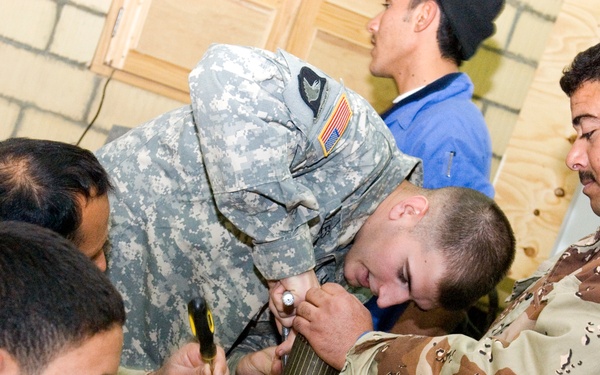 The image shows a joint military training session where a U.S. Army soldier in ACU (Army Combat Uniform) is working alongside several soldiers wearing desert camouflage uniforms, likely Iraqi Security Forces. They are gathered around a workbench collaboratively handling what appears to be a mechanical or ordnance component. The setting appears to be an indoor training facility with cinder block and plywood walls.