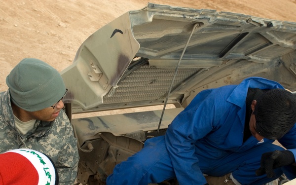 Three individuals are gathered around an open hood of a military vehicle, likely a HMMWV (Humvee), performing or observing mechanical work. One person is wearing US Army combat uniform (ACU pattern) with a gray beanie and glasses, while another in a blue mechanic's coverall is leaning into the engine compartment. The setting appears to be an arid, desert environment.