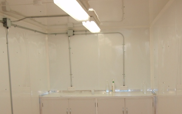 The image shows the interior of a clean, white-walled modular or portable room, likely a laboratory or clinical workspace. It features built-in white base cabinets with a work surface along the back wall, overhead fluorescent lighting, and exposed conduit runs along the ceiling and walls. The space appears newly installed or recently set up, with minimal items on the countertop.