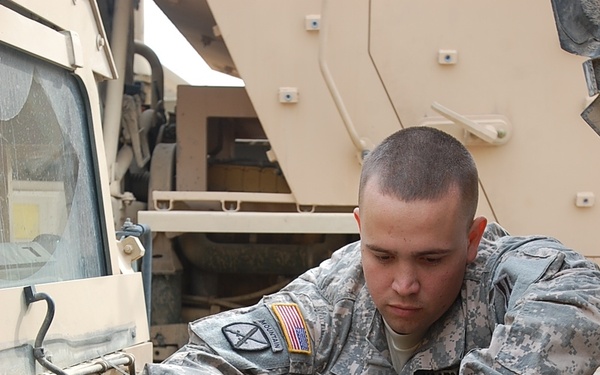 A U.S. Army soldier named Flaherty, wearing Army Combat Uniform (ACU) with a 10th Mountain Division patch and U.S. flag patch, is performing maintenance on a military vehicle engine. The soldier is leaning over an open engine compartment, working with both hands on engine components. The vehicle appears to be a tan/desert-colored military tactical vehicle, likely a HUMVEE or similar platform.