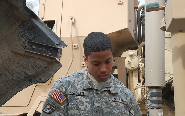 A U.S. Army soldier identified by name tape as 'Joseph' is performing maintenance work on a military vehicle, likely a MRAP or similar tactical wheeled vehicle. The soldier is wearing ACU (Army Combat Uniform) with digital camouflage pattern and black mechanic's gloves, indicating hands-on mechanical work. The setting appears to be a forward operating base or motor pool in a desert environment.