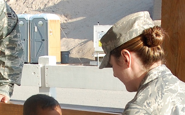 A female U.S. Air Force service member in ABU (Airman Battle Uniform) is giving a high-five to a young smiling boy. The interaction appears warm and joyful, with the child laughing enthusiastically. The setting appears to be an outdoor military base or construction area.