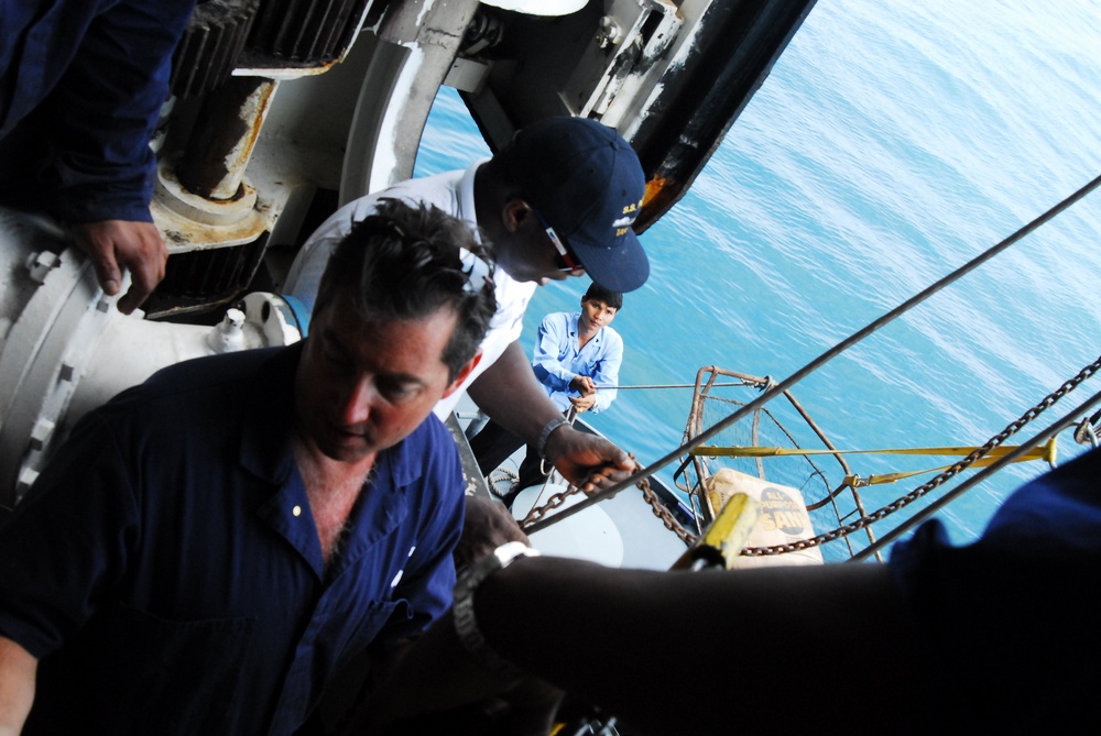 The image shows several individuals working on the deck or exterior of a naval or maritime vessel, with open ocean visible in the background. Workers appear to be handling chains, ropes, and rigging equipment on what looks like a ship's exterior structure. The scene suggests a maritime maintenance or boarding operation in progress.