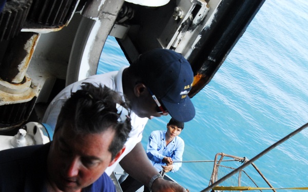 The image shows several individuals working on the deck or exterior of a naval or maritime vessel, with open ocean visible in the background. Workers appear to be handling chains, ropes, and rigging equipment on what looks like a ship's exterior structure. The scene suggests a maritime maintenance or boarding operation in progress.