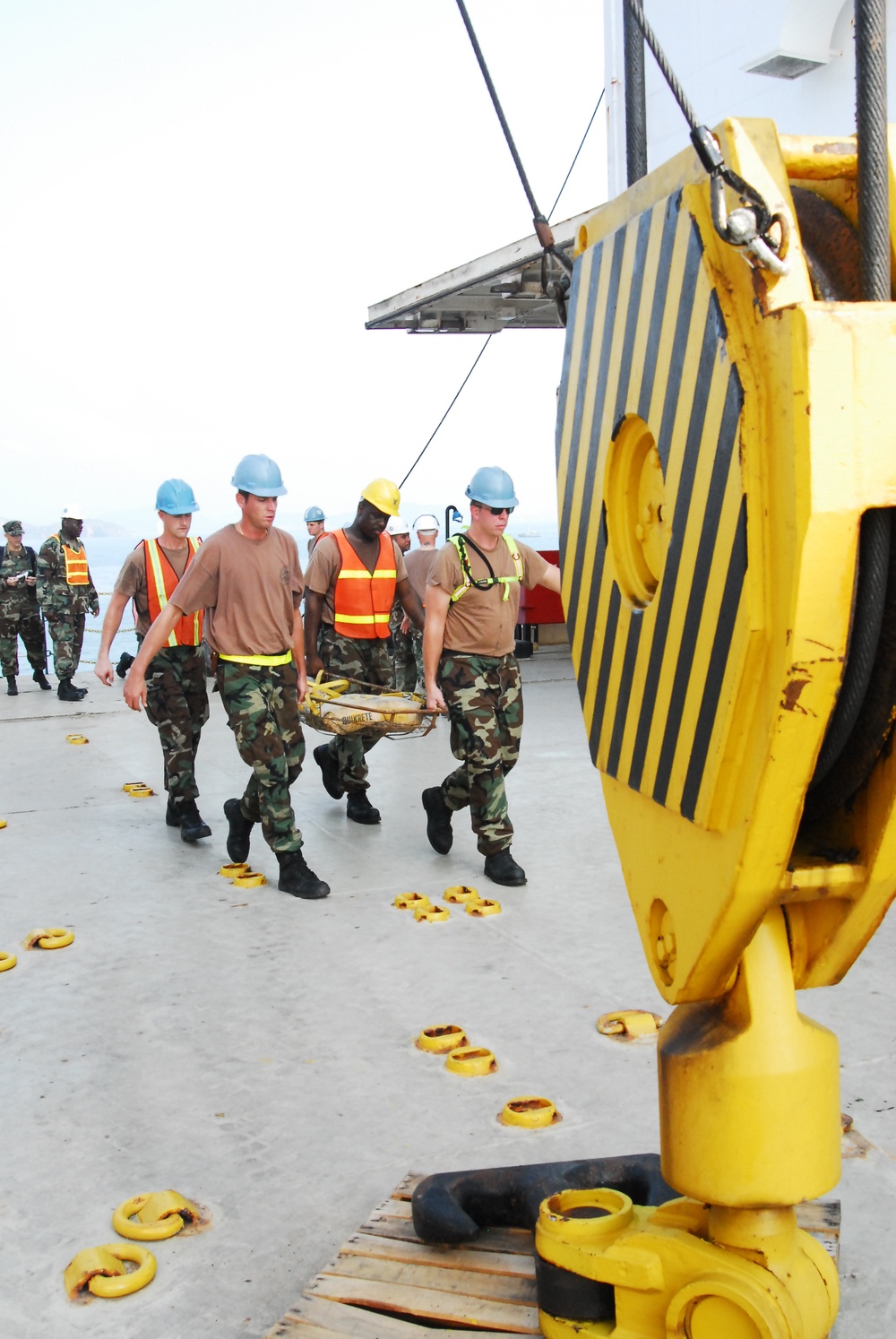 Military personnel in camouflage uniforms and safety vests are carrying a stretcher or litter across what appears to be the deck of a large vessel or pier. Several individuals wear hard hats in blue and yellow, along with high-visibility orange safety vests. A large yellow crane hook and rigging equipment is prominently visible in the foreground.