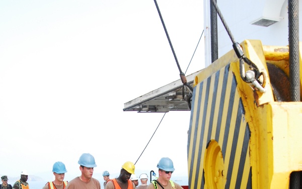 Military personnel in camouflage uniforms and safety vests are carrying a stretcher or litter across what appears to be the deck of a large vessel or pier. Several individuals wear hard hats in blue and yellow, along with high-visibility orange safety vests. A large yellow crane hook and rigging equipment is prominently visible in the foreground.