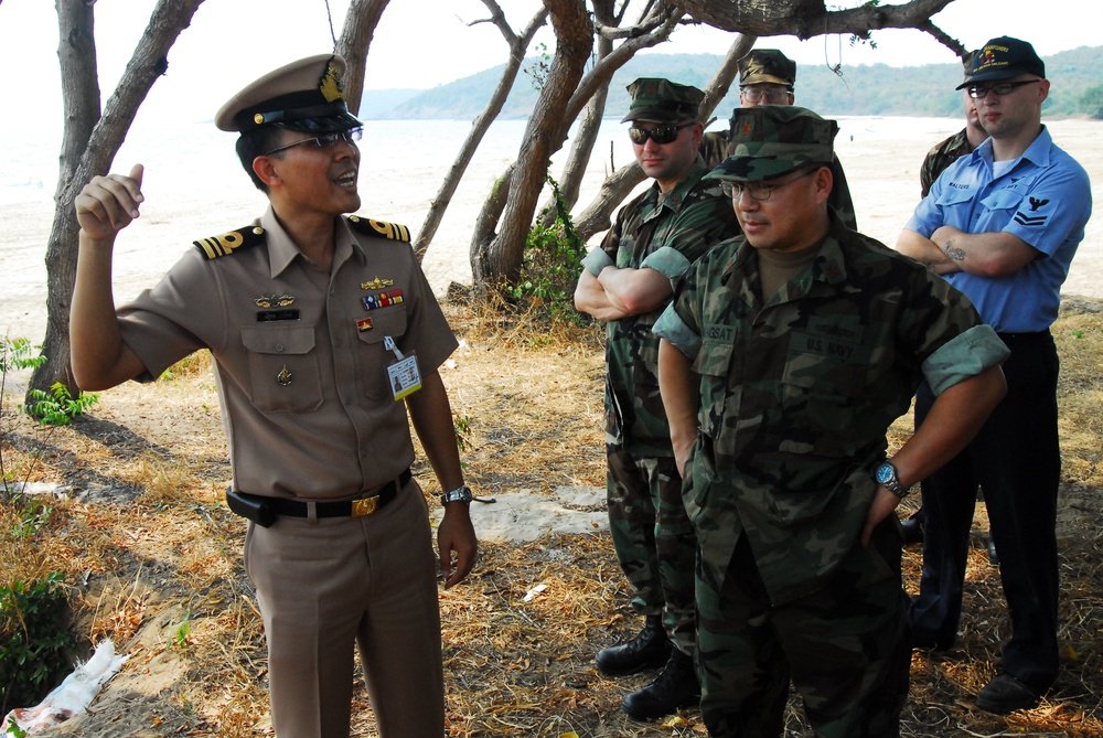 A group of military personnel from what appears to be multiple nations are gathered outdoors near a beach or coastal area. A senior officer in a tan/khaki uniform with a white peaked cap and ribbons appears to be briefing or gesturing to a group that includes individuals in woodland camouflage uniforms and one person in a light blue uniform. The setting suggests a joint military exercise, liaison meeting, or site survey.