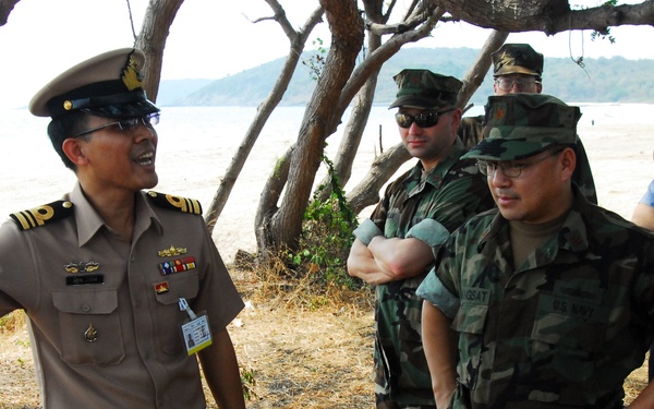 A group of military personnel from what appears to be multiple nations are gathered outdoors near a beach or coastal area. A senior officer in a tan/khaki uniform with a white peaked cap and ribbons appears to be briefing or gesturing to a group that includes individuals in woodland camouflage uniforms and one person in a light blue uniform. The setting suggests a joint military exercise, liaison meeting, or site survey.