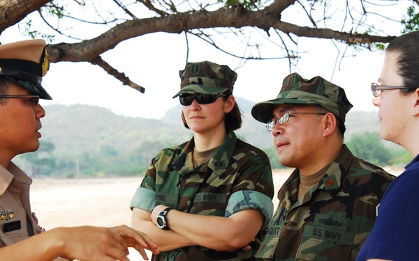 Four individuals are engaged in a conversation outdoors in an arid, open area beneath a tree. One person wears a tan/khaki military dress uniform with a peaked cap, two others wear U.S. Navy woodland camouflage uniforms, and one civilian wears a blue shirt. The scene appears to be a military coordination or briefing meeting.