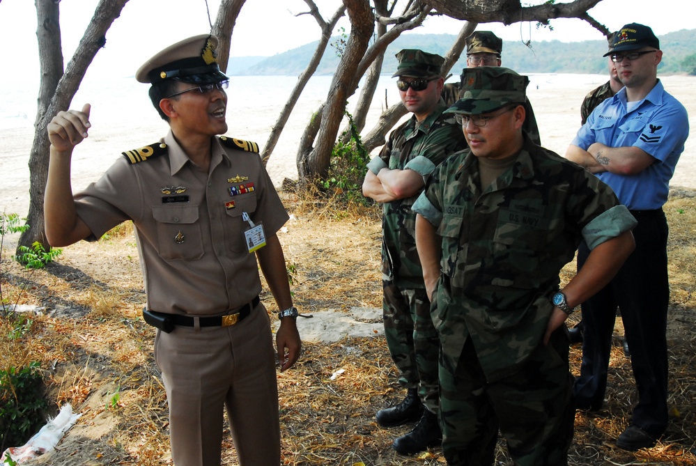 A group of military personnel are gathered outdoors near a beach and tree line. A senior officer in a tan/khaki uniform with a peaked cap and ribbons appears to be briefing or addressing several others in woodland camouflage uniforms and one individual in a light blue uniform. The setting suggests a multinational military exercise or coordination meeting.