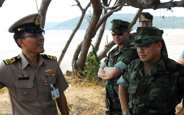 A group of military personnel are gathered outdoors near a beach and tree line. A senior officer in a tan/khaki uniform with a peaked cap and ribbons appears to be briefing or addressing several others in woodland camouflage uniforms and one individual in a light blue uniform. The setting suggests a multinational military exercise or coordination meeting.