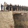 A large group of men, the majority wearing black uniforms with tactical boots, are running in formation across a deteriorating stone bridge or elevated road structure in an arid, desert-like environment. One individual at the front is dressed in civilian clothing (beige tracksuit and white sneakers), suggesting a leadership or instructor role. The setting and uniforms indicate this is likely a physical training exercise for a security or paramilitary force.