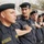A group of uniformed personnel from the 'Oil Protection Force' are standing in formation, with each person placing their hand on the shoulder of the person in front of them. They are wearing dark navy/black uniforms with black berets bearing a badge insignia. The setting appears to be an outdoor training or parade ground with trees visible in the background.