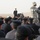 An outdoor briefing or training session is taking place in an arid, desert-like environment. A U.S. military soldier in ACU (Army Combat Uniform) and a civilian or contractor in dark clothing are addressing a large group of uniformed personnel seated on the ground. The seated personnel appear to be local security forces or police, wearing dark blue/black uniforms with caps.