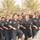 A large group of men in dark navy blue uniforms are running in formation while chanting or shouting, suggesting a military or police training exercise. They wear matching uniforms with visible patches and insignia, indicating an organized security force. A person in camouflage military uniform is visible in the foreground right, observing the formation.